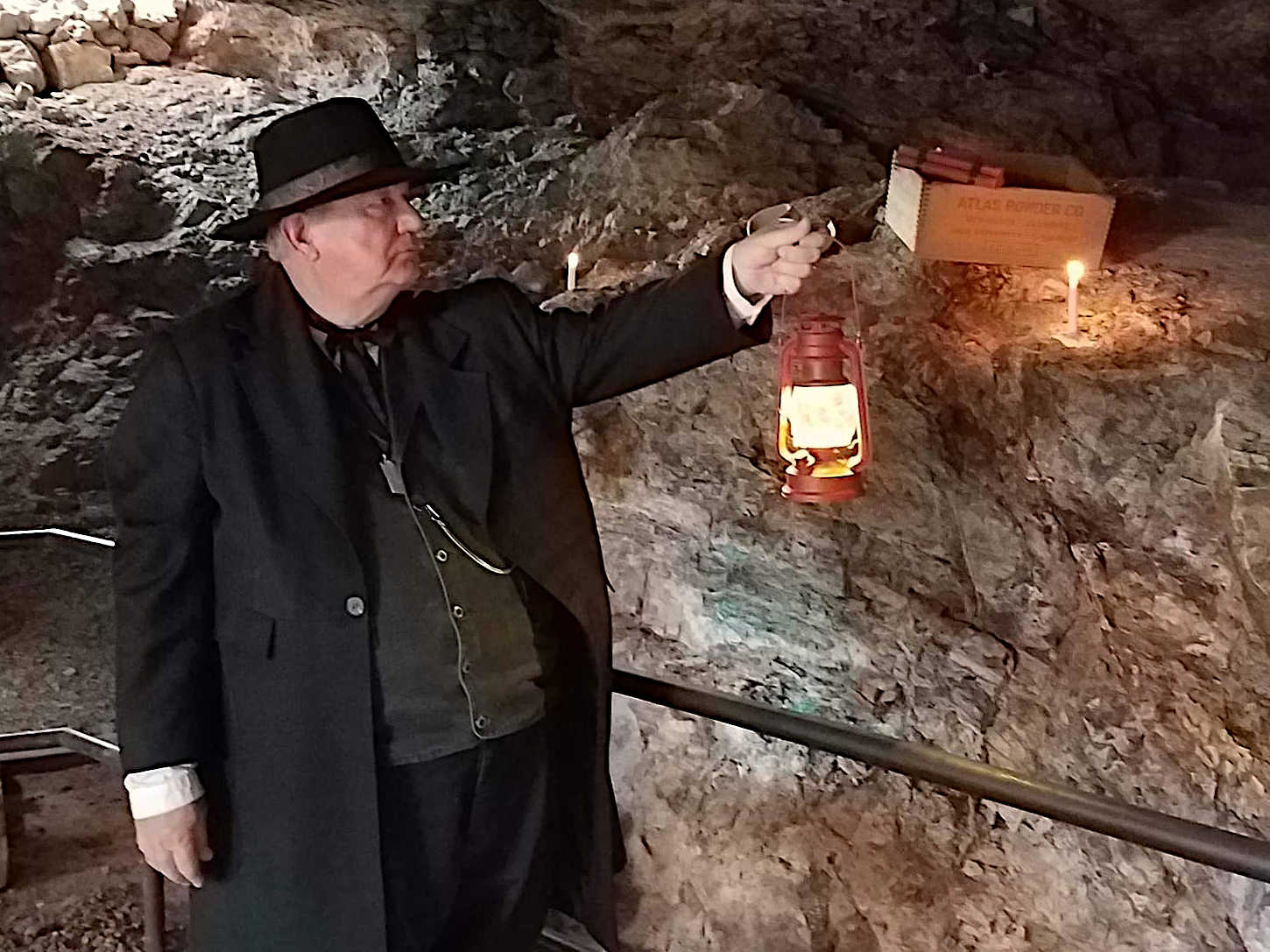 A man in period clothing holding a lit lantern inside a dimly lit mine, with a wooden box labeled 'Atlas Powder Co' and a candle on a rocky ledge in the background.