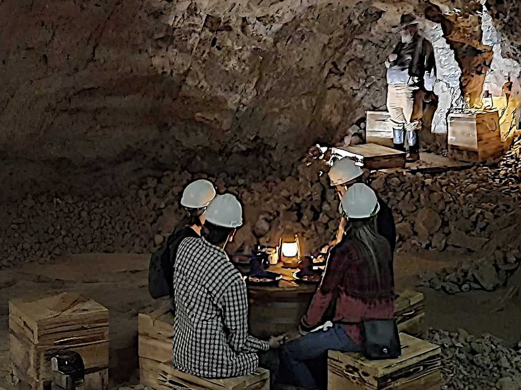 Group of visitors wearing hard hats sitting at wooden tables inside a dimly lit mine, listening to an actor dressed in period clothing standing on a rocky ledge. Lanterns provide light in the underground setting.