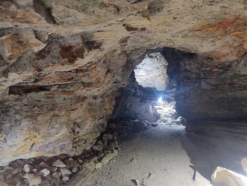 Underground mine tunnel with rugged rock walls, scattered debris, and light streaming in from an opening in the distance.