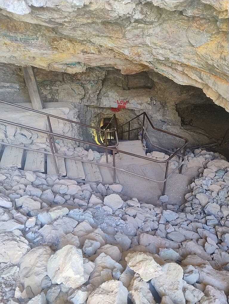 Entrance to an underground mine with a wooden walkway and metal handrails, surrounded by large rocks and rugged stone walls.