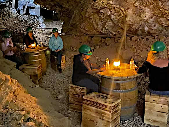 Visitors in hard hats sitting at barrel tables inside a mine, illuminated by lanterns, surrounded by rocky walls and wooden crates.