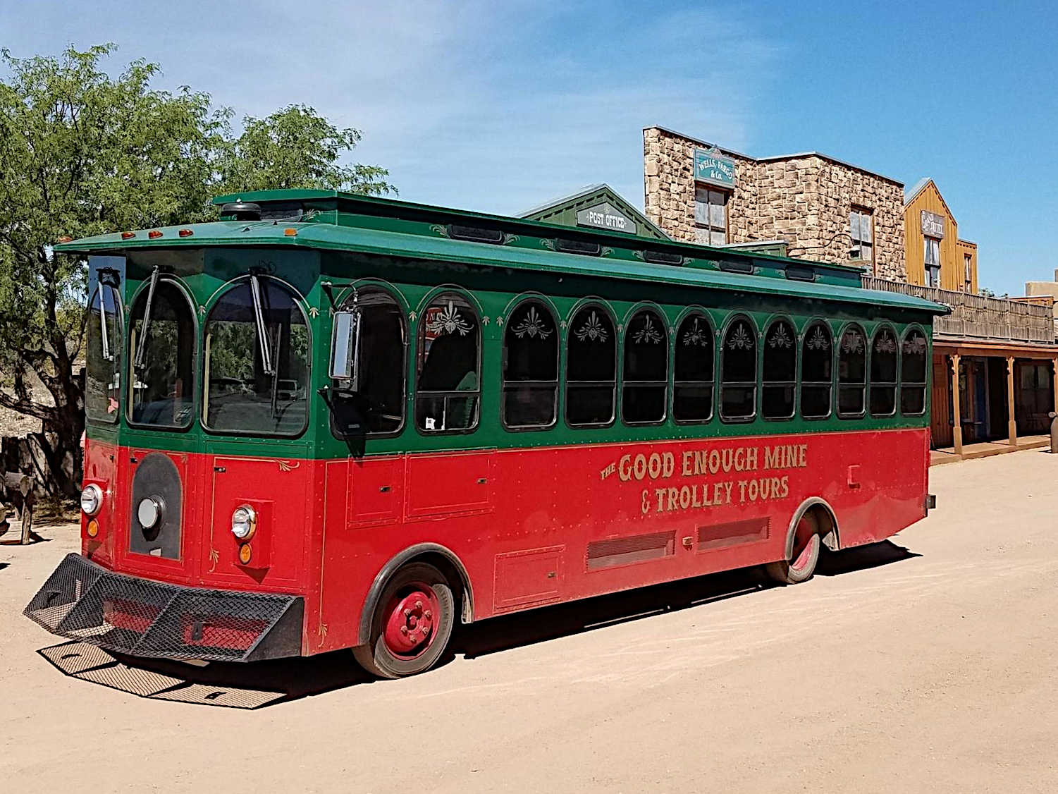 A red and green trolley bus labeled 'The Good Enough Mine & Trolley Tours' parked on a dirt road with old western-style buildings and desert trees in the background.